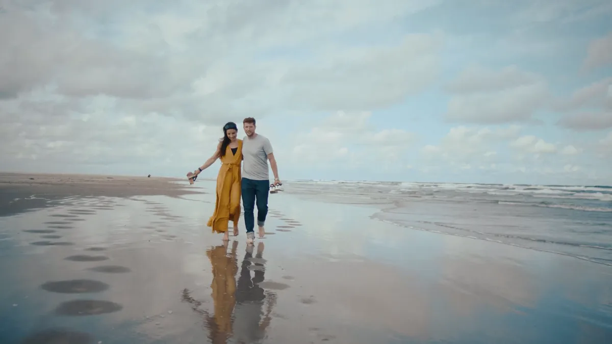 Standbild aus dem Borkum-Werbespot: ein Paar geht bei Ebbe über den weiten, nassen Strand, dahinter Wolkenhimmel und Nordsee.