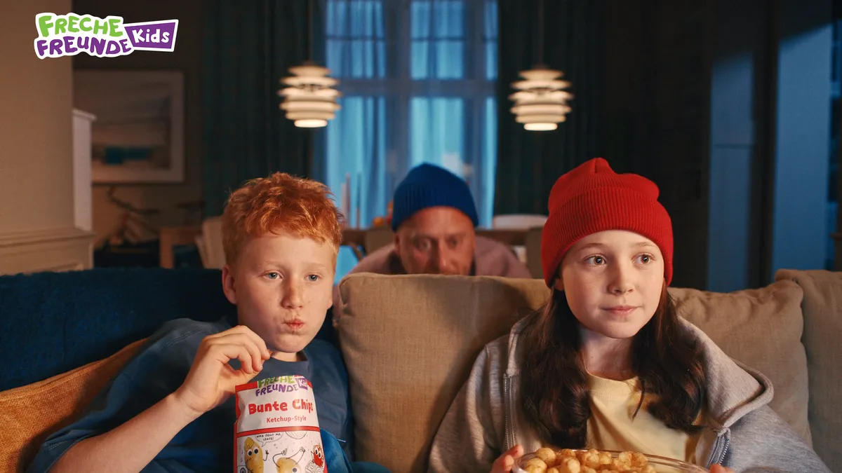 Standbild aus dem Freche-Freunde-Werbespot: zwei Kinder auf einem Sofa greifen nach einer Schale Snacks, dahinter lugt ein Erwachsener hervor.
