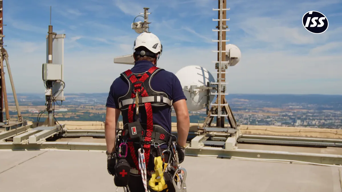 Standbild aus dem ISS-Recruiting-Film: ein Techniker mit Helm und Sicherungsgurt auf einem Dach zwischen Antennen, im Hintergrund eine weite Landschaft.