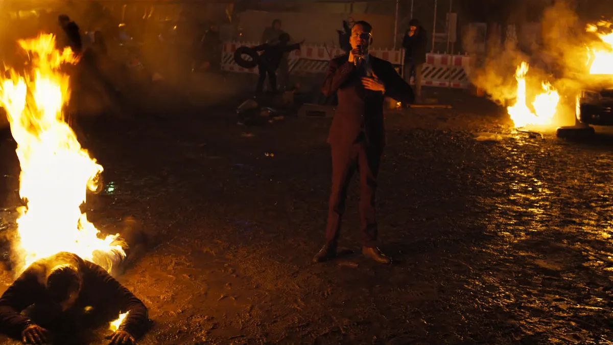 Standbild aus dem K.I.Z-Musikvideo: ein brennender Mann am Boden, dahinter ein Redner mit Mikrofon und brennende Autos.