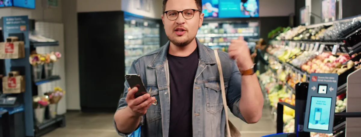 Standbild aus dem REWE-Film: der Host steht mit dem Handy in einem REWE-Markt, im Hintergrund die Regale und das Pick-&-Go-Logo.