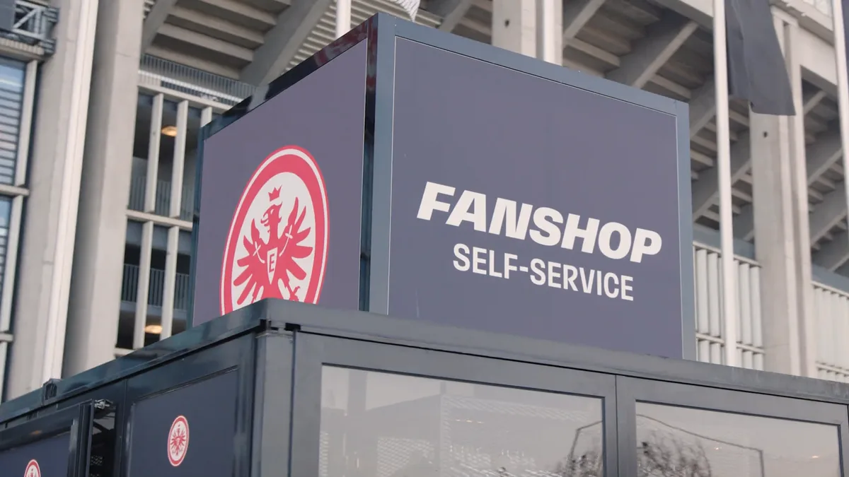 Standbild aus dem VR-Payment-Film: der Fanshop-Self-Service-Container mit Eintracht-Frankfurt-Wappen vor der Stadionfassade.